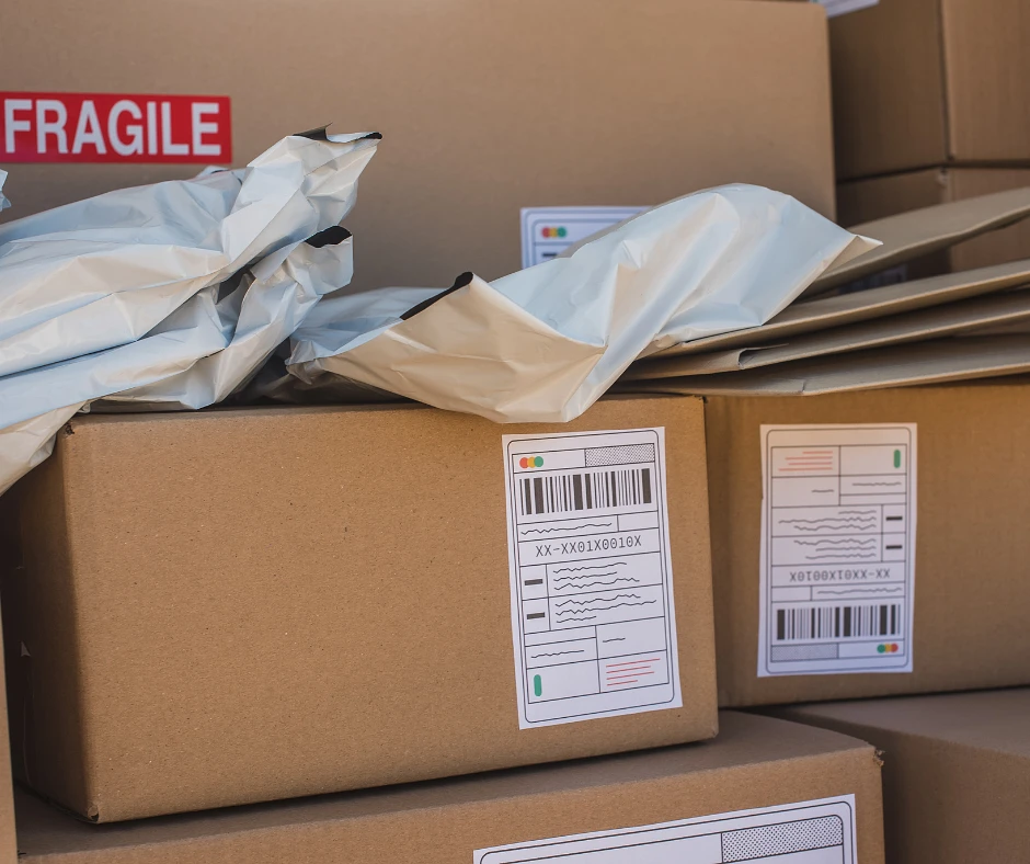 A stack of cardboard boxes labeled with barcodes and a "fragile" sign, surrounded by crumpled shipping envelopes.