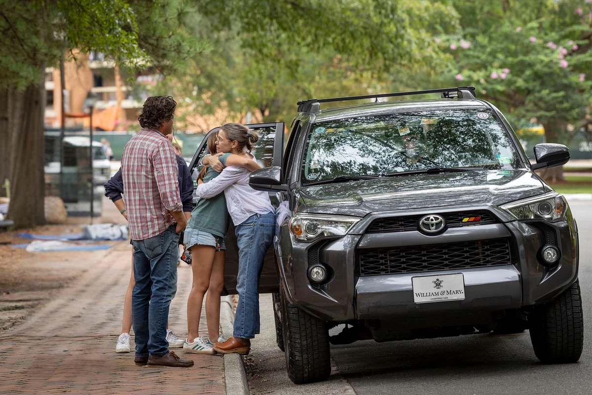 A group of people is standing near an SUV, with one person hugging another on a tree-lined street.