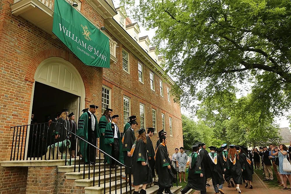 Graduates in caps and gowns are walking outside a historic building under a "William & Mary" banner.