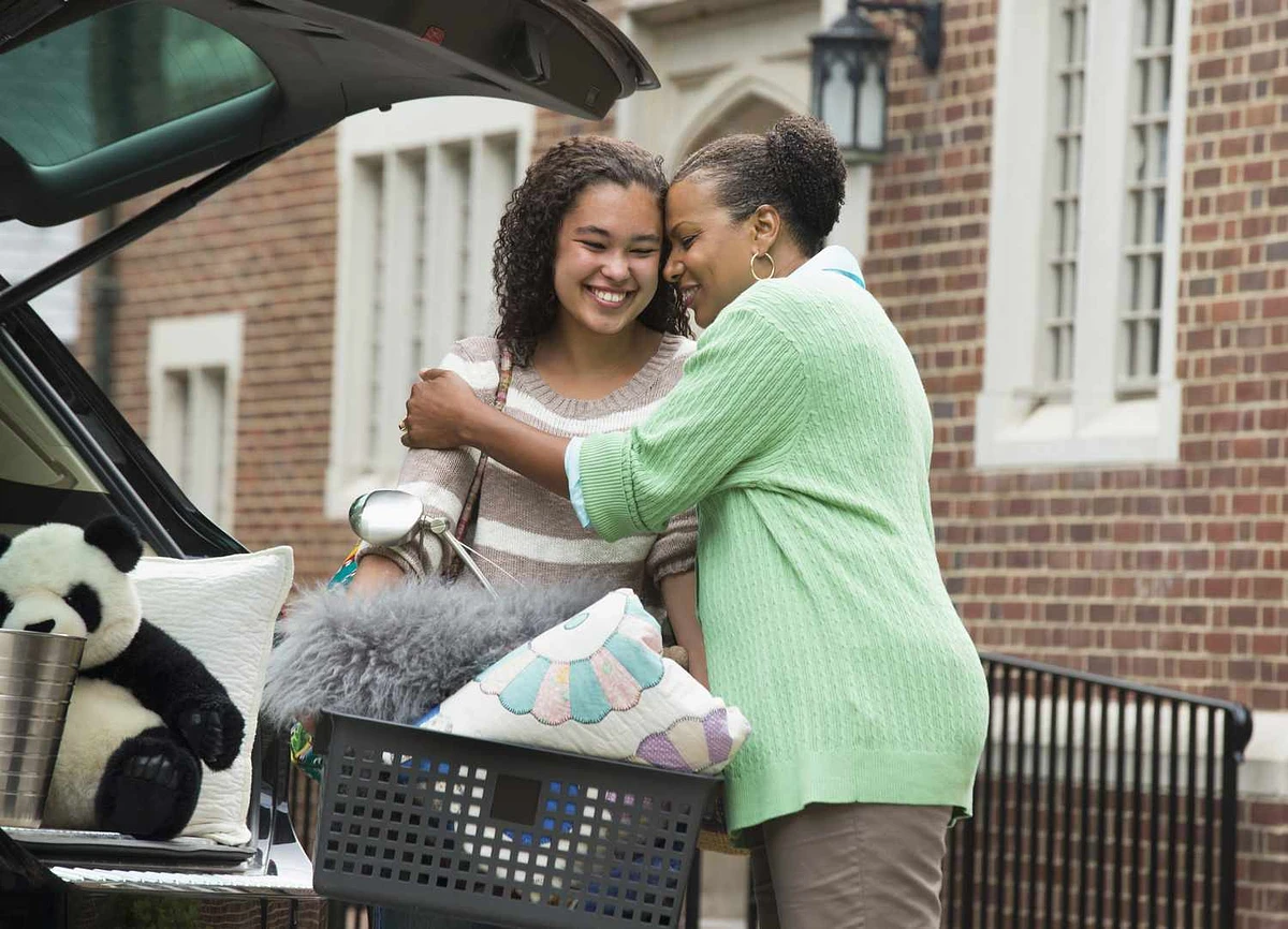 A mother and daughter share a warm hug while unloading belongings from a car in front of a brick building.